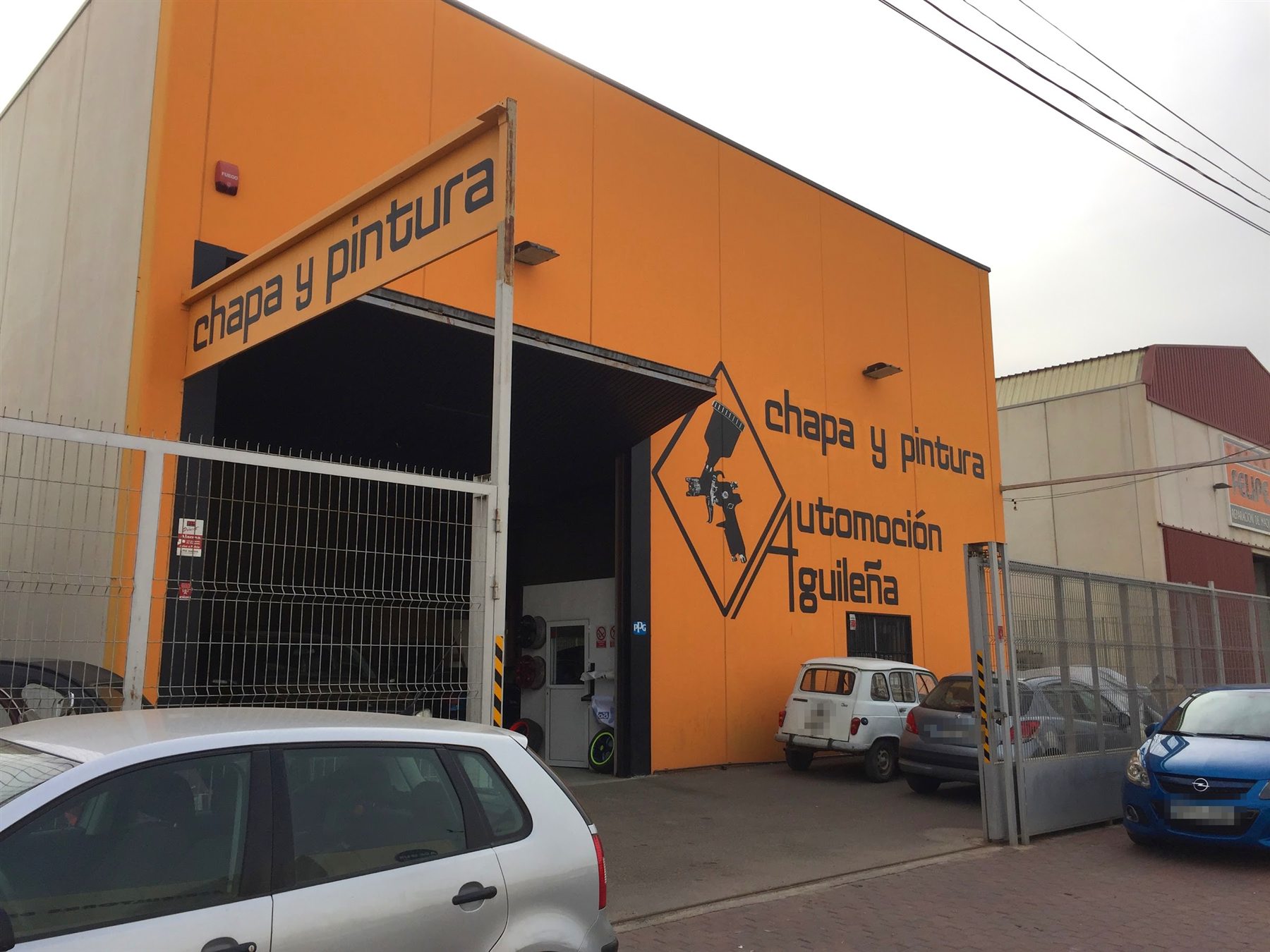 Fachada naranja de la nave con el rótulo de chapa y pintura Automoción Aguileña pintado en el muro